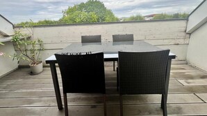 Una terraza en la azotea con una mesa de vidrio y dos sillas de mimbre, una planta en maceta y vistas a árboles y edificios al fondo.