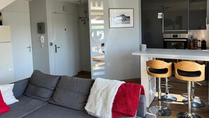 Un moderno salón con un sofá gris, una manta roja y una manta blanca y esponjosa. Se observa una zona de cocina con encimera blanca y electrodomésticos de acero inoxidable. Hay dos taburetes de madera a la vista.