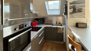 Una cocina moderna con electrodomésticos de acero inoxidable, horno empotrado, microondas y una tostadora roja. Hay una ventana sobre el fregadero, una silla de madera y un cuadro en la pared.