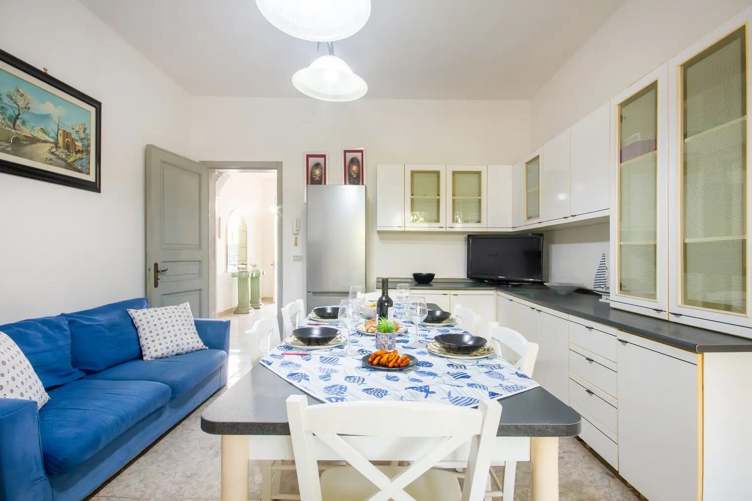 Una cocina compacta con armarios blancos, una mesa de comedor dispuesta con platos, tazones y una botella. En la zona de estar, hay un sofá azul con cojines estampados.
