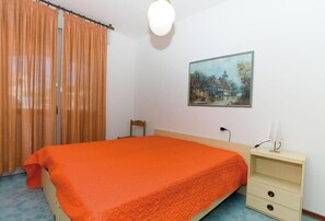 A bedroom with a bed, a nightstand, a chair, a painting on the wall, and a window with curtains. - Terraced House in a Residence With a Pool (San Michele al Tagliamento)