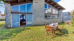 A stone house with large windows, a patio with wooden furniture, and a visible interior with a fireplace. - Charmante Maison proche plage (Saint-Pierre-d'Oléron)