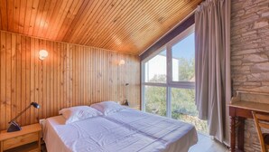 A bedroom with a wooden ceiling, a large bed with white bedding, a nightstand with a lamp, and a stone wall. - Charmante Maison proche plage (Saint-Pierre-d'Oléron)