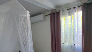A bedroom with a bed, a nightstand, a window with curtains, and a mosquito net. - Le lotus rental in terre sainte, 2 minutes from the beach (Saint-Pierre)