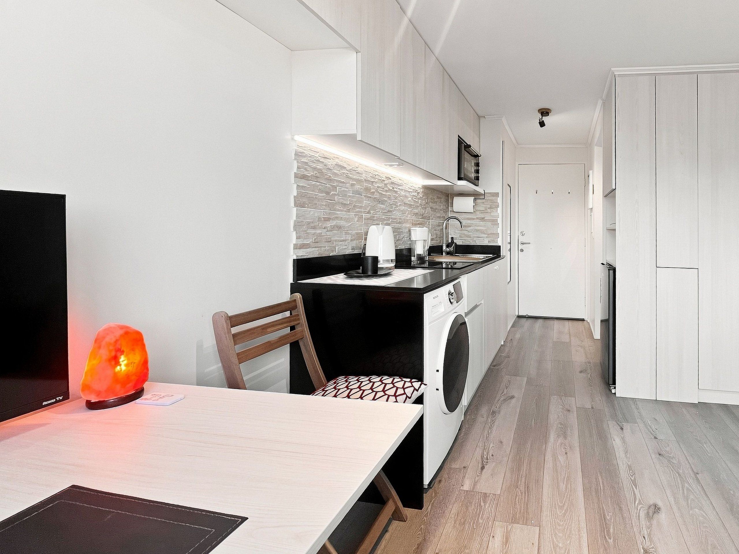 A compact kitchen with a washing machine, a dining area with a table and chairs, and a TV on a stand.