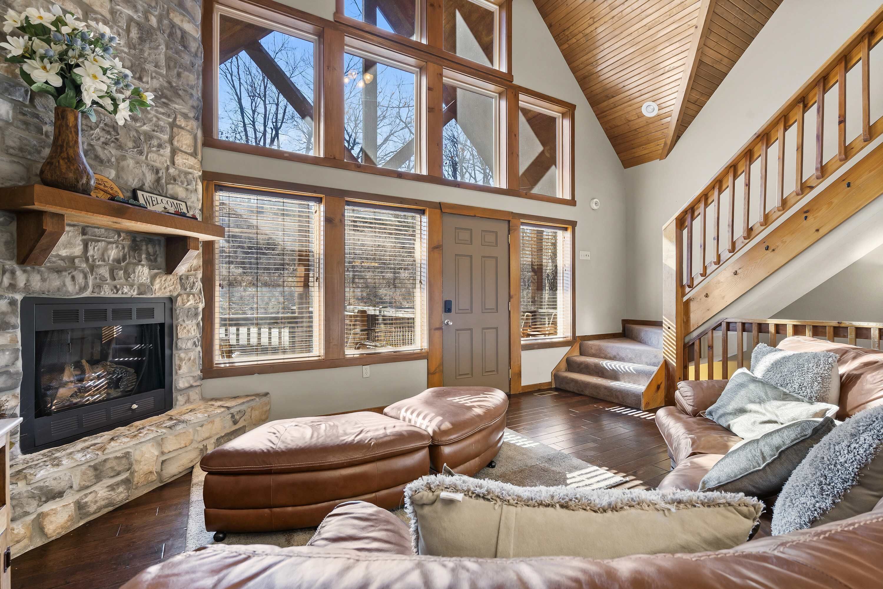 A spacious living room with a stone fireplace, leather seating, large windows, and a wooden staircase.