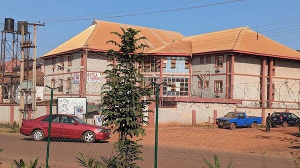 Un bâtiment devant lequel est garée une voiture rouge, avec une voiture bleue en arrière-plan, et une clôture en treillis métallique qui entoure la propriété.