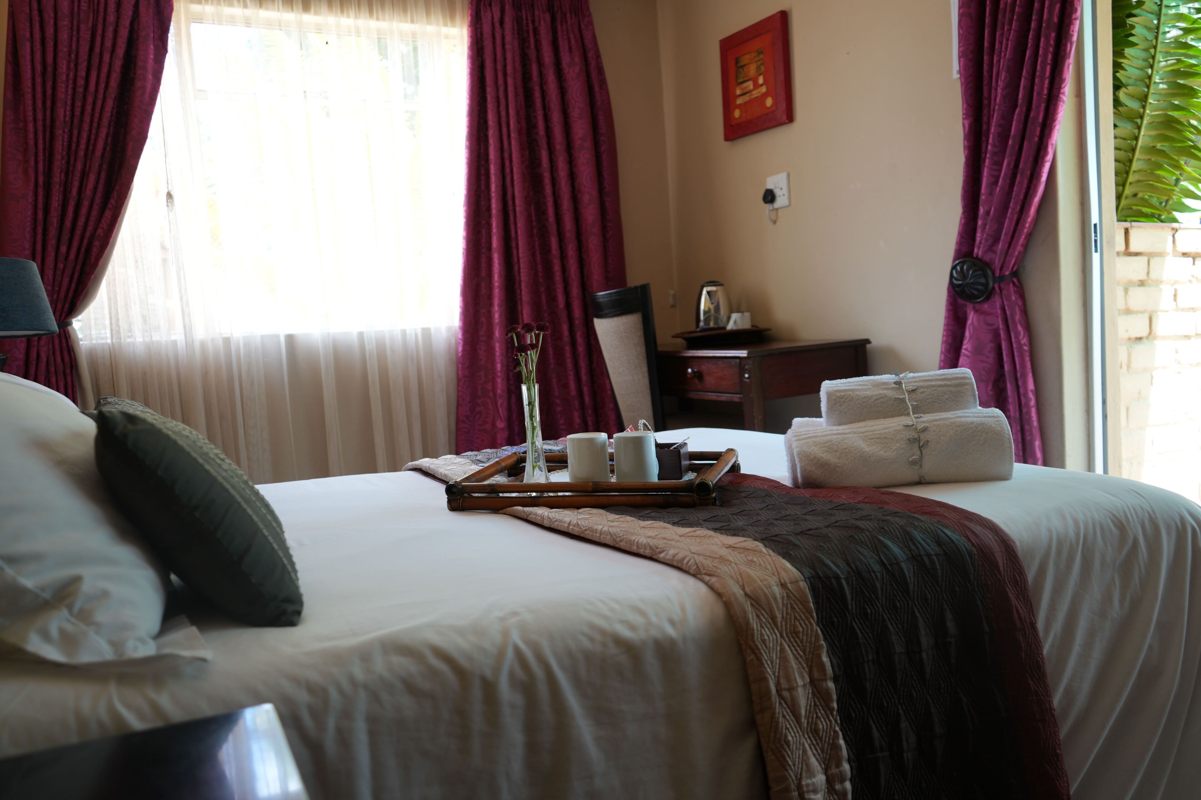 A hotel room with a bed, pillows, a tray with a vase, and a window with curtains.