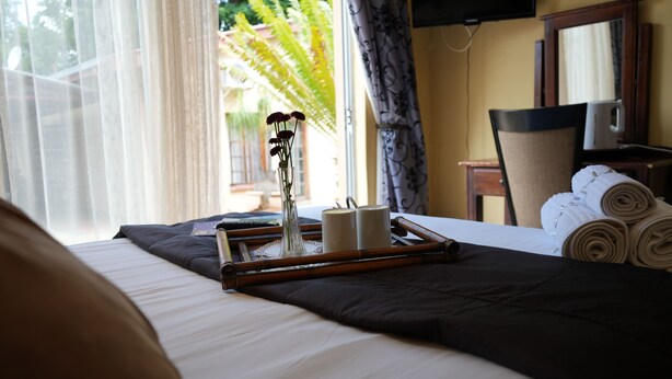Una cama bien tendida con un recipiente de artículos de aseo, un jarrón con flores y vistas a una palmera a través de la ventana.
