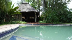 Una piscina con borde de azulejos, rodeada de exuberante vegetación y una estructura con techo de paja.