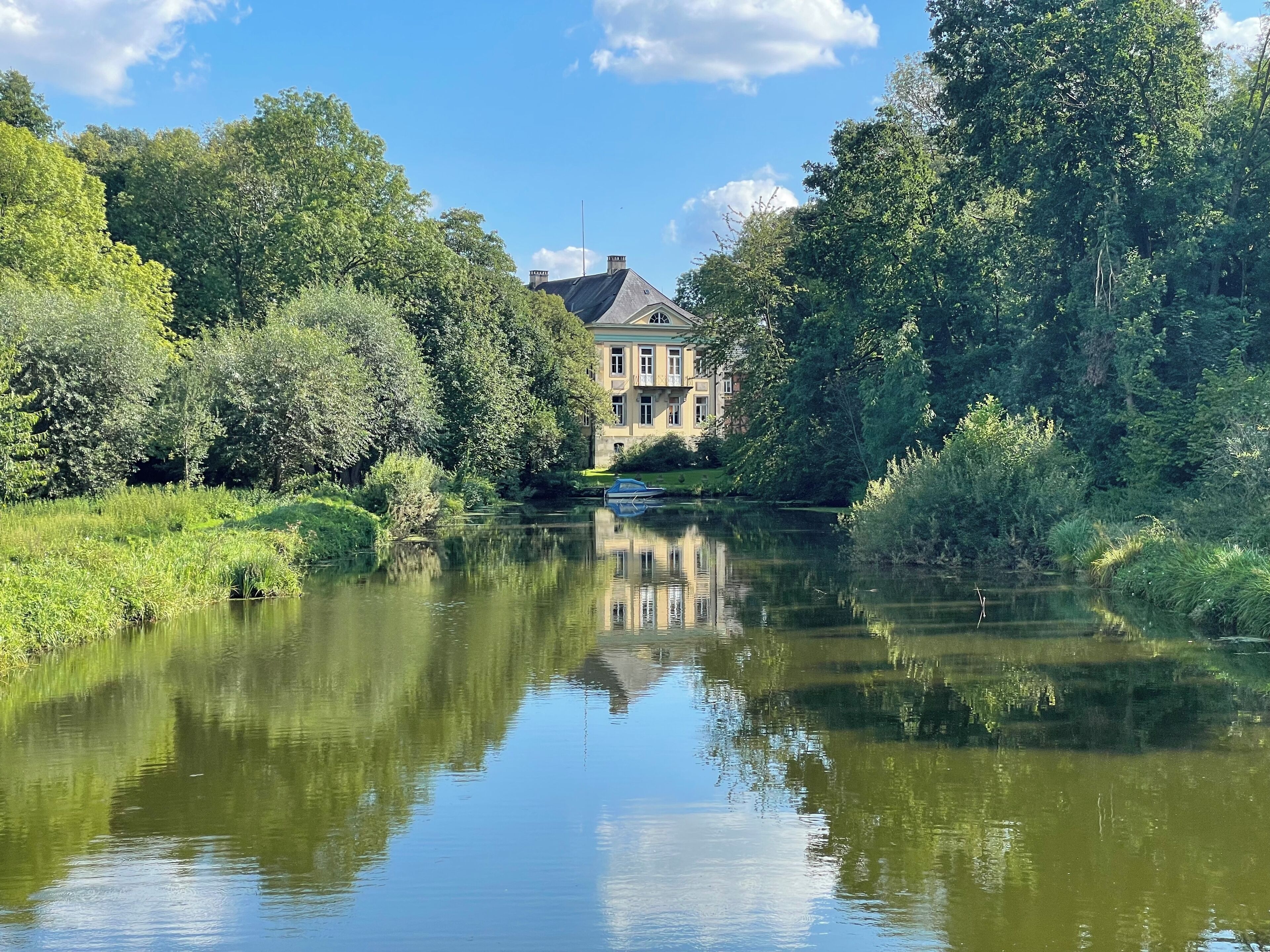 Eine friedliche Flusslandschaft mit einem historischen Gebäude, das sich im Wasser spiegelt, umgeben von üppigem Grün.
