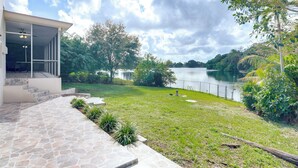 A modern house with a stone patio leading to a covered outdoor area, a grassy lawn, and a lake view. - 8 Mi to Wynwood: Waterfront Retreat w/ Lanai (North Miami)