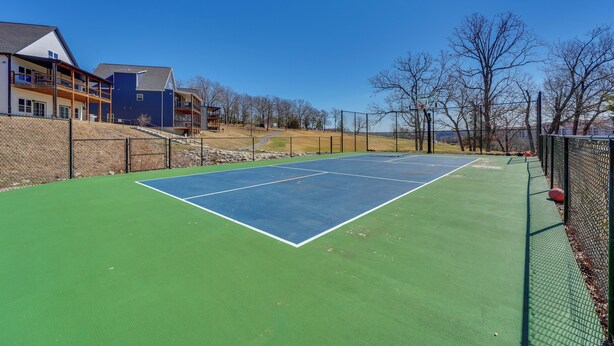 Una cancha de tenis exterior con superficie verde, rodeada por una valla y adyacente a un edificio con balcones.