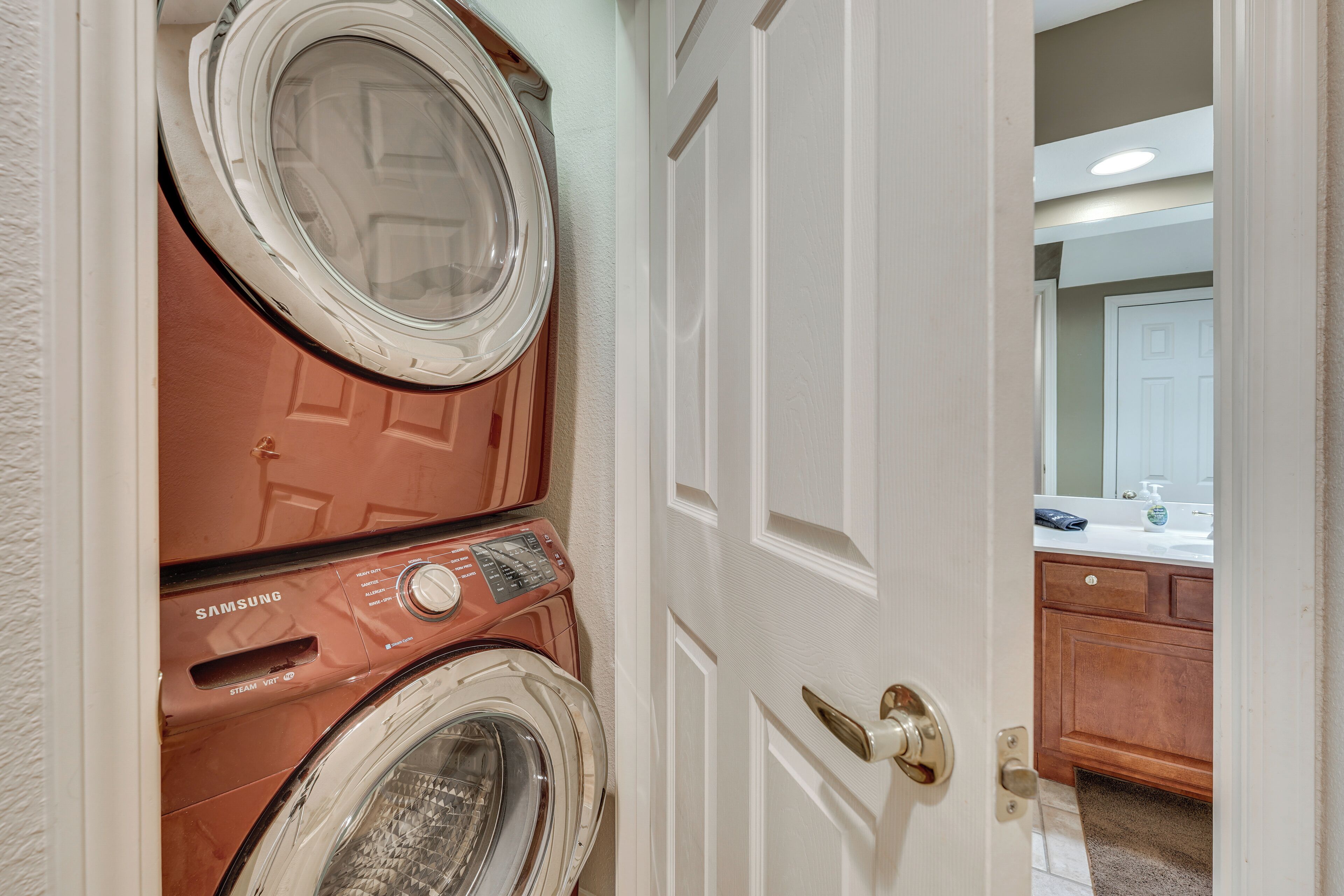 Une buanderie équipée d’un lave-linge et d’un sèche-linge Samsung, avec une porte blanche et une vue sur une salle de bain comprenant un lavabo et un miroir.