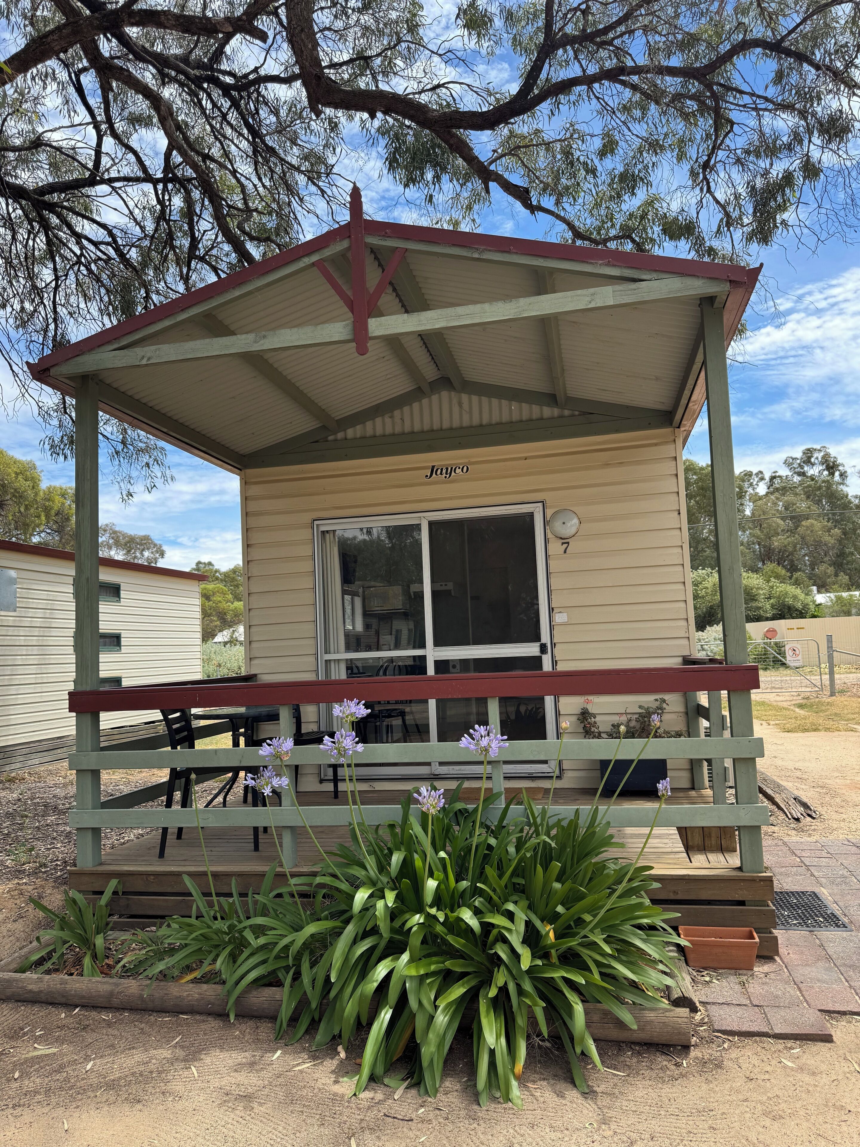 A small, single-story structure with a red roof and beige walls, labeled "Jayco" with the number 7. It features a wooden deck with a bench and a table, surrounded by green plants and purple flowers.