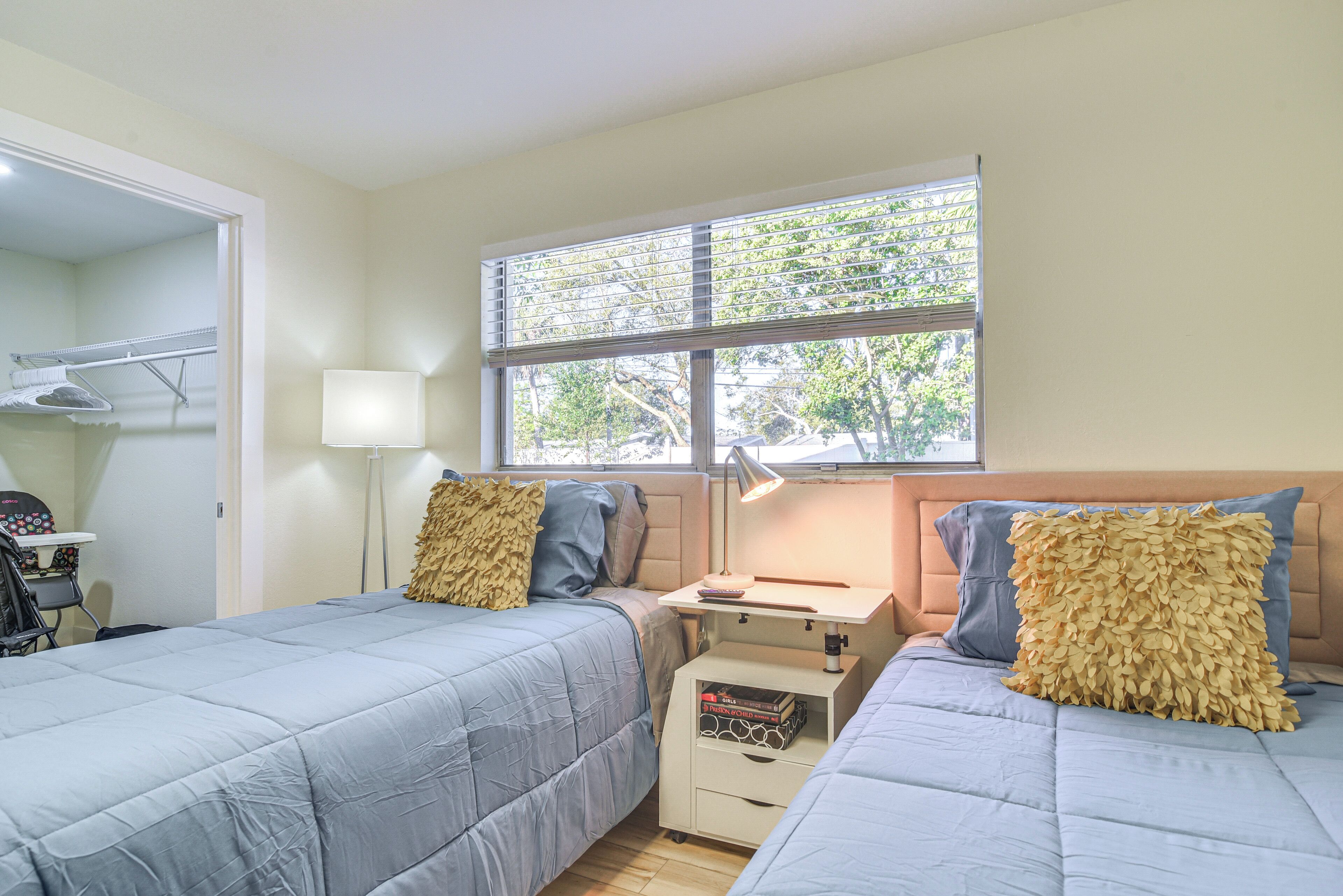 Une chambre avec deux lits, une table de chevet avec une lampe, une fenêtre avec des stores et un petit bureau avec une chaise.