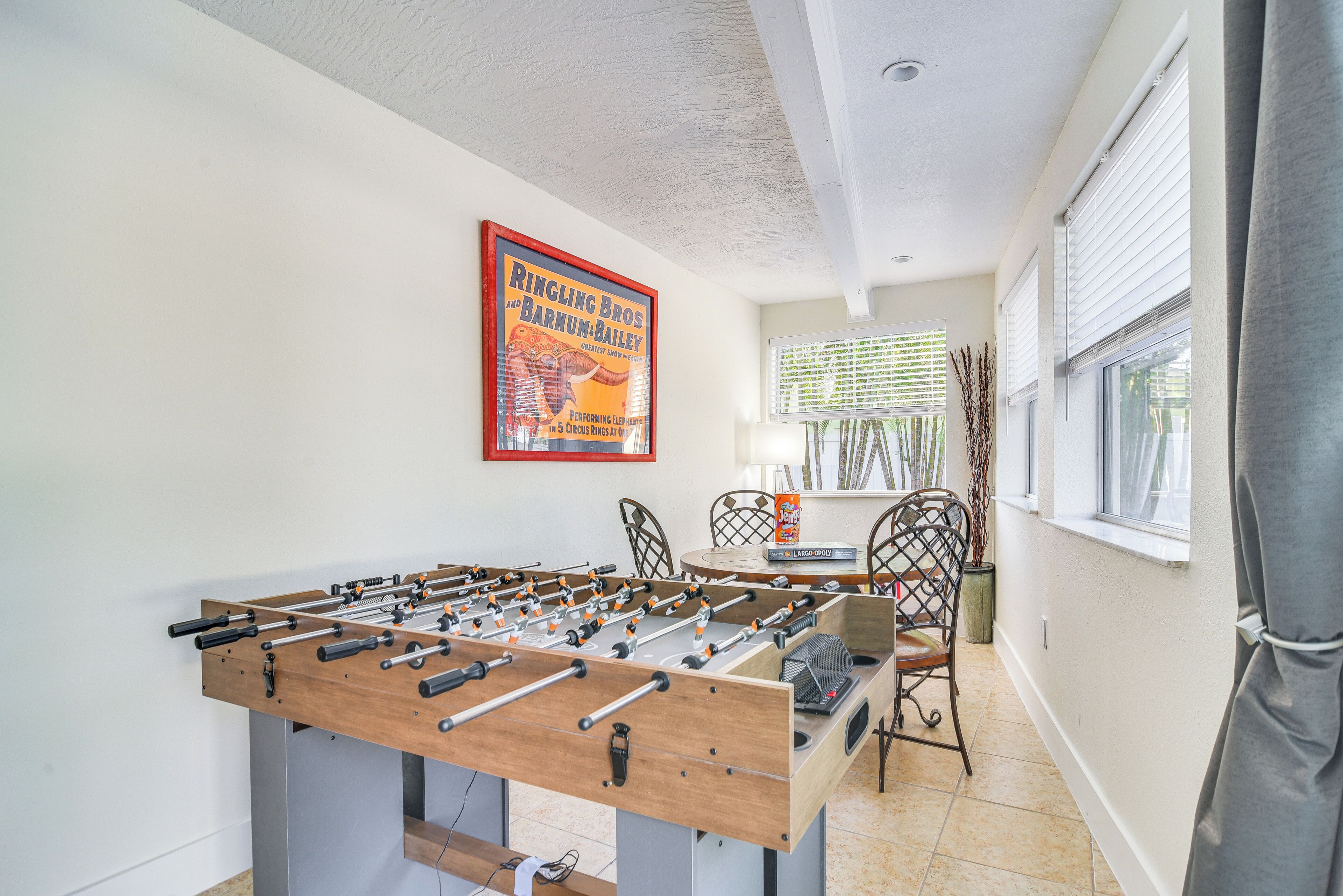 A foosball table in a room with a poster on the wall, a window with blinds, and a chair.