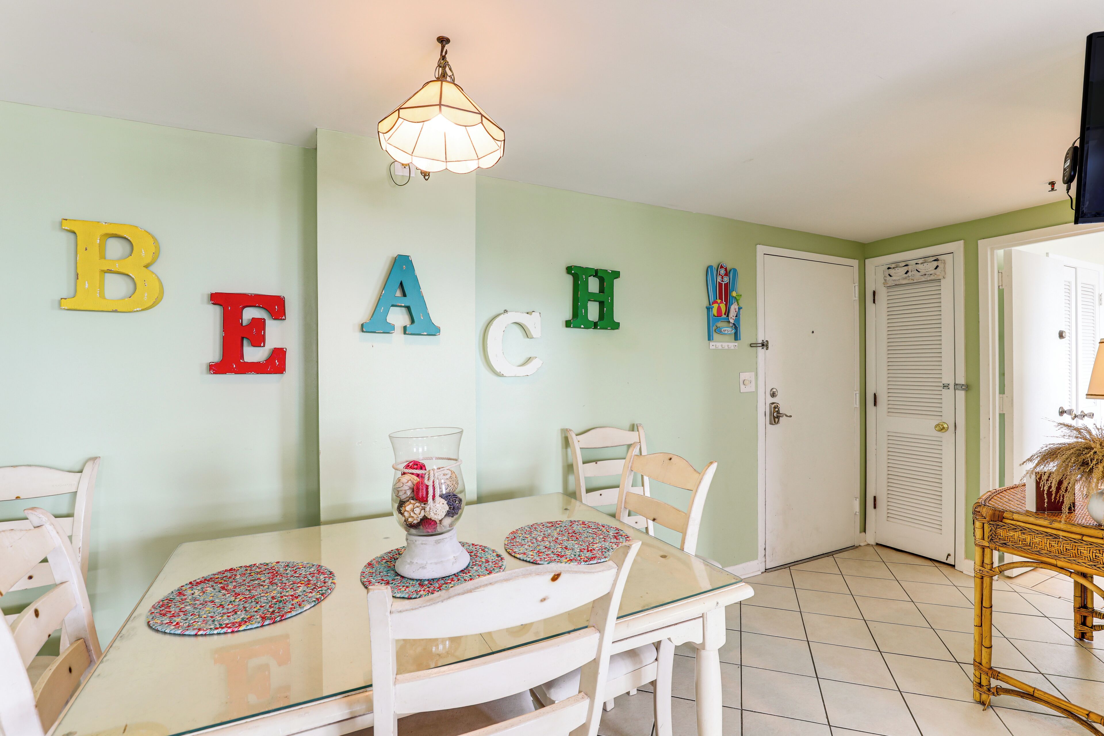 A dining area with a glass table, white chairs, and a wall with colorful letters spelling "BEACH".