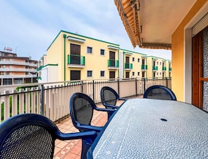 A balcony with a tiled table and chairs, overlooking a row of yellow buildings with green balconies. - Modern Apartment With a Spacious Terrace (San Michele al Tagliamento)