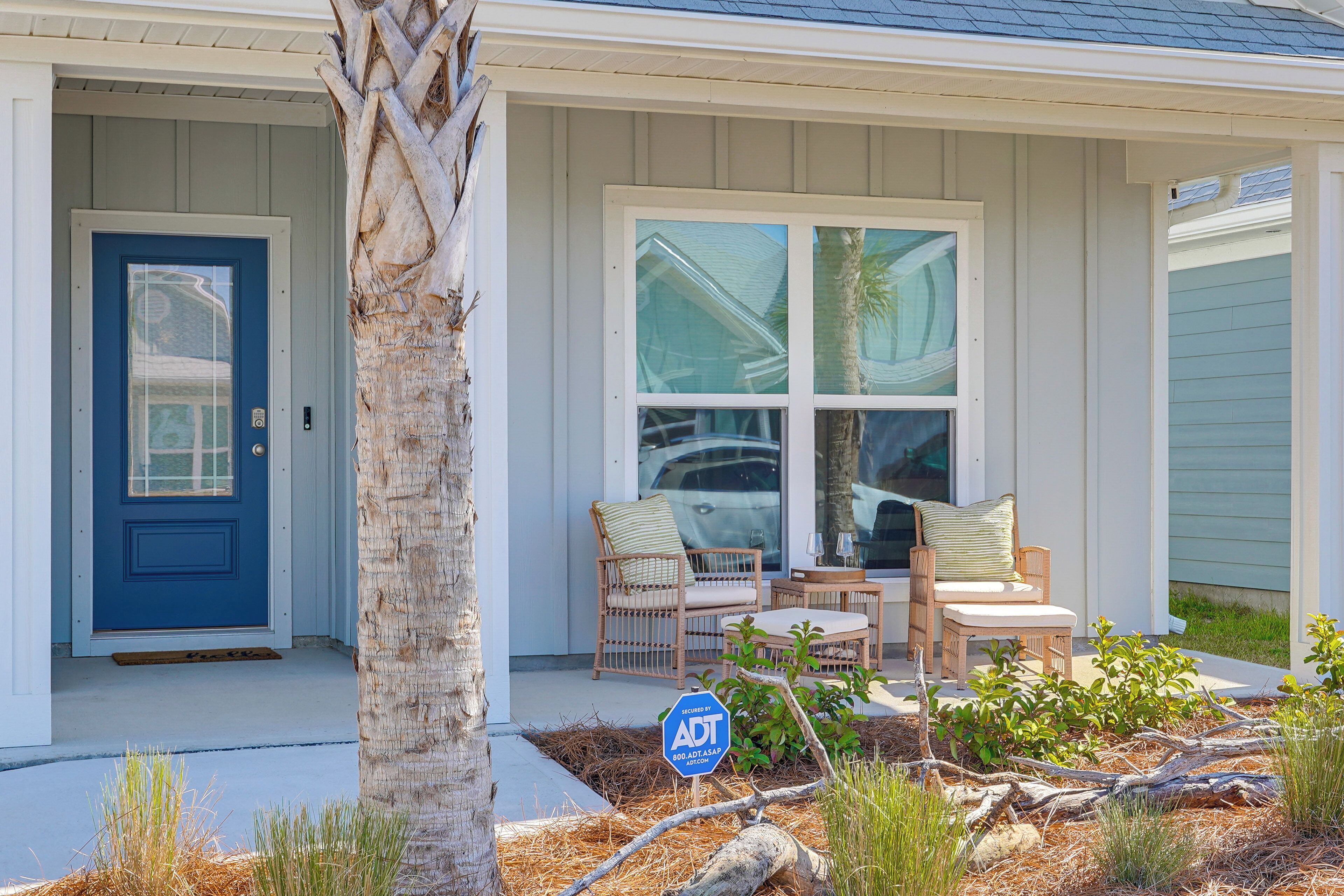 A house with a blue door, two wicker chairs with cushions, and a small garden area with a sign.