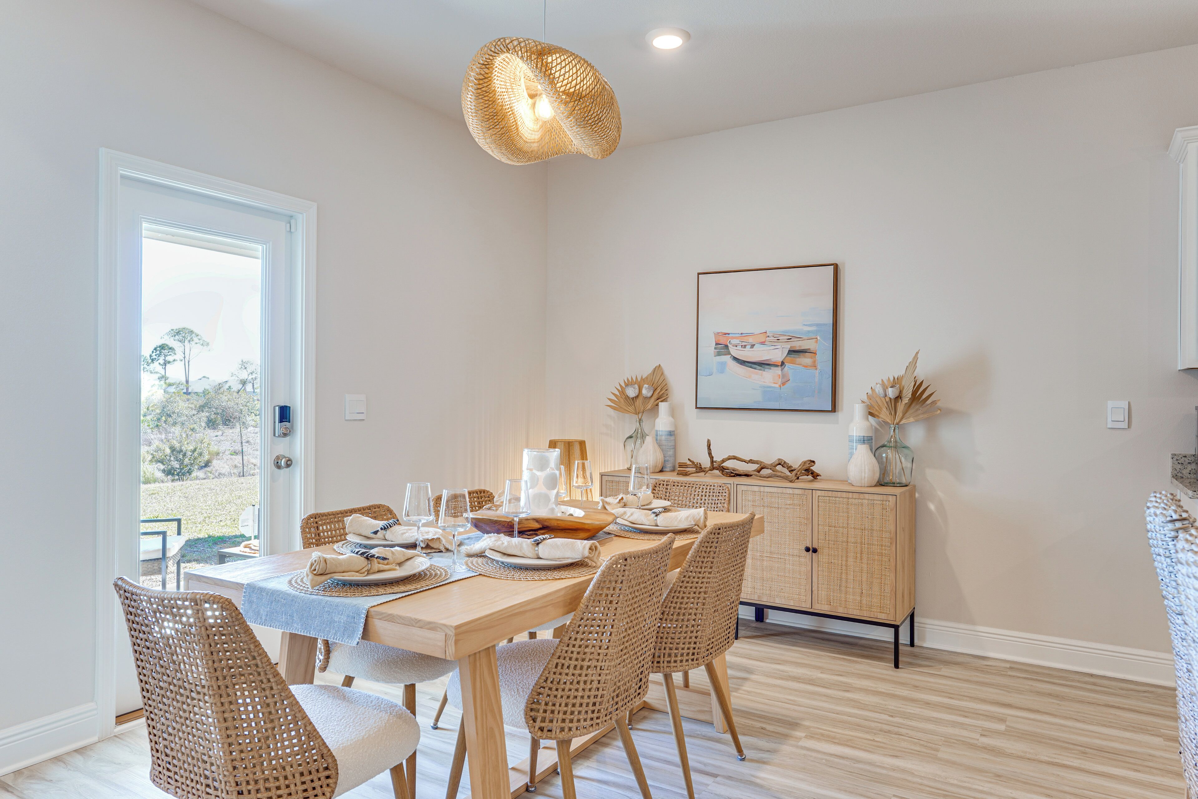 A dining area with a wooden table set for six, surrounded by wicker chairs, a wicker cabinet, and a hanging light fixture.