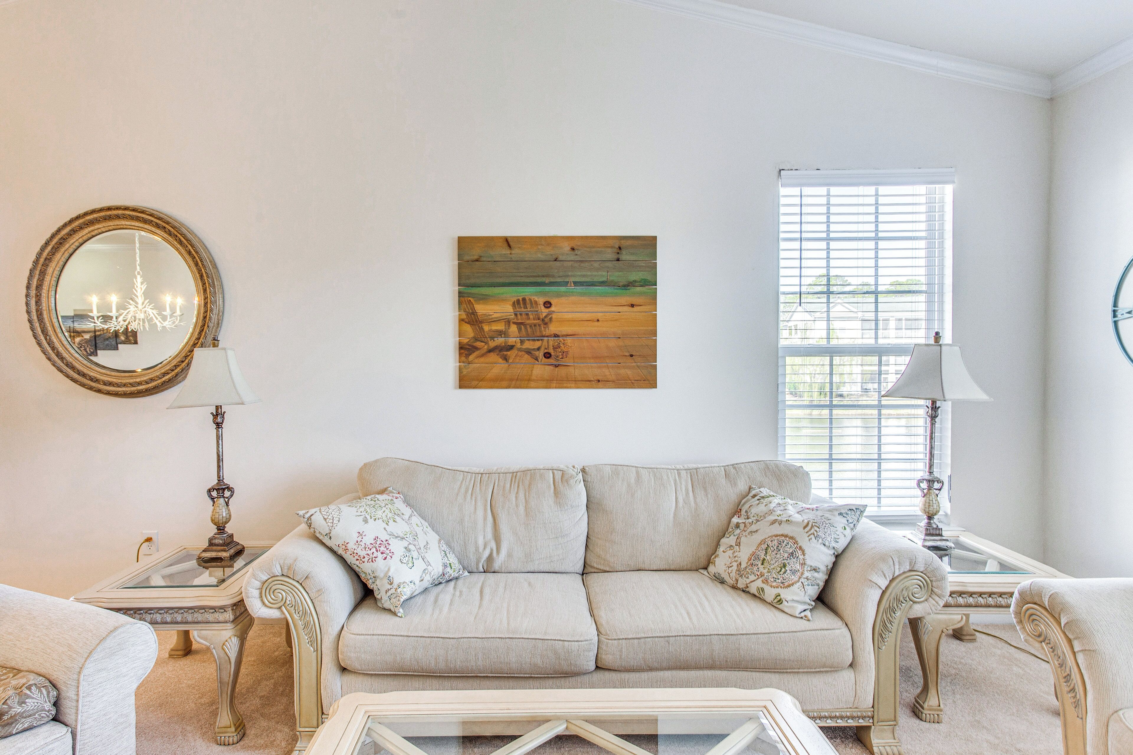 A living room with a beige sofa, two side tables, a glass coffee table, a painting, a chandelier, and a window with blinds.