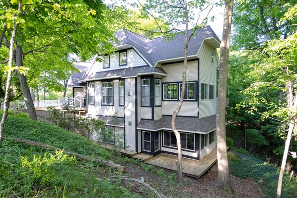 A two-story house with a large deck, surrounded by dense greenery and trees.
