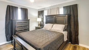 A bedroom with a bed, dark wooden dresser, nightstand with lamp, ceiling fan, and window with curtains. - Near Shops & Eats: Home w/ Large Yard in Waycross! (Waycross)
