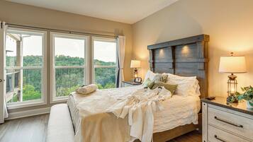 A bedroom with a large bed, two bedside lamps, a nightstand, and a view of a forest through the windows.