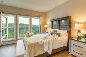 A bedroom with a large bed, two bedside lamps, a nightstand, and a view of a forest through the windows. - Luxury Cabin, Mountain Views, Hot Tub, EV Charger (Ellijay)