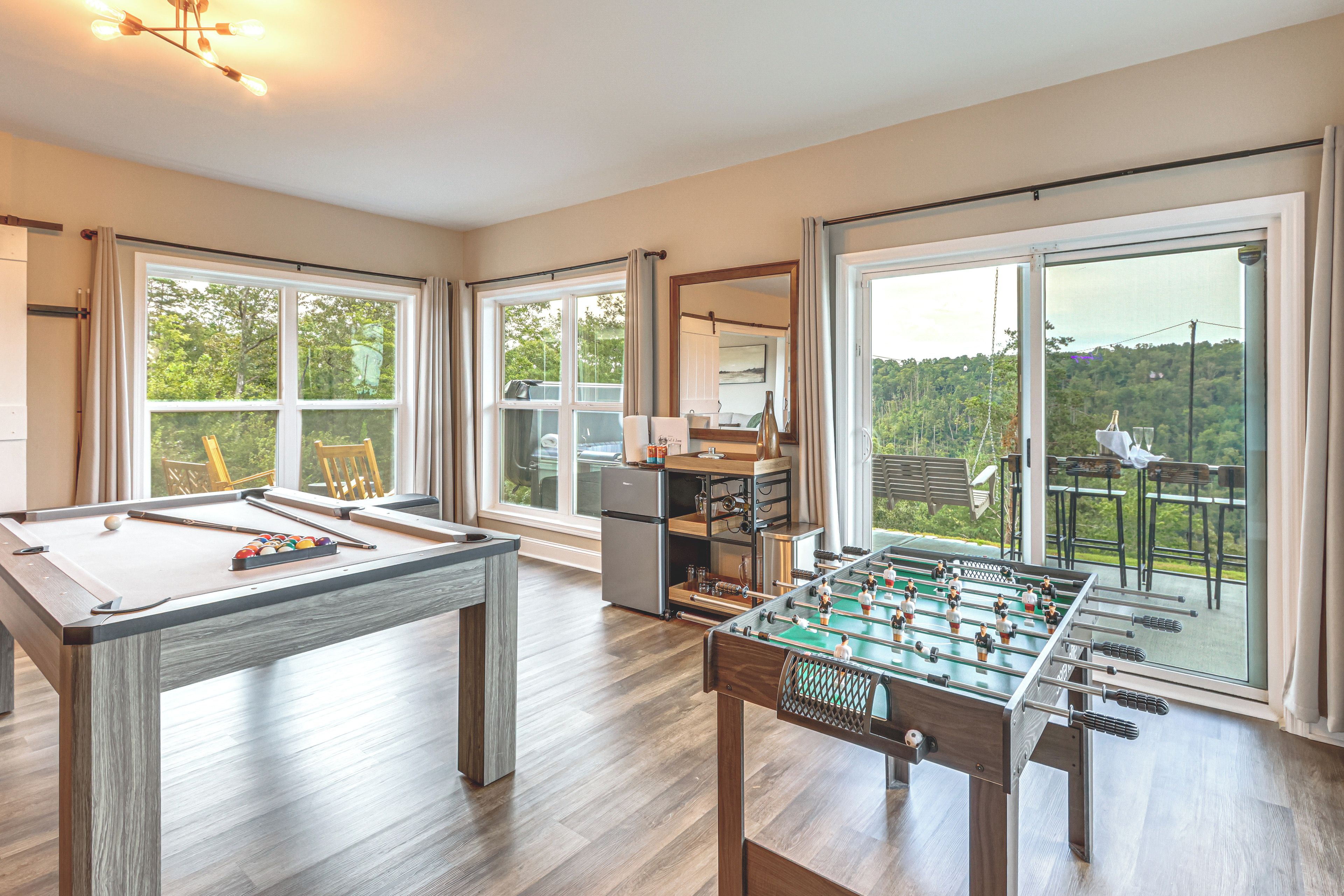 A room with a pool table, foosball table, and a large window with a view of greenery.