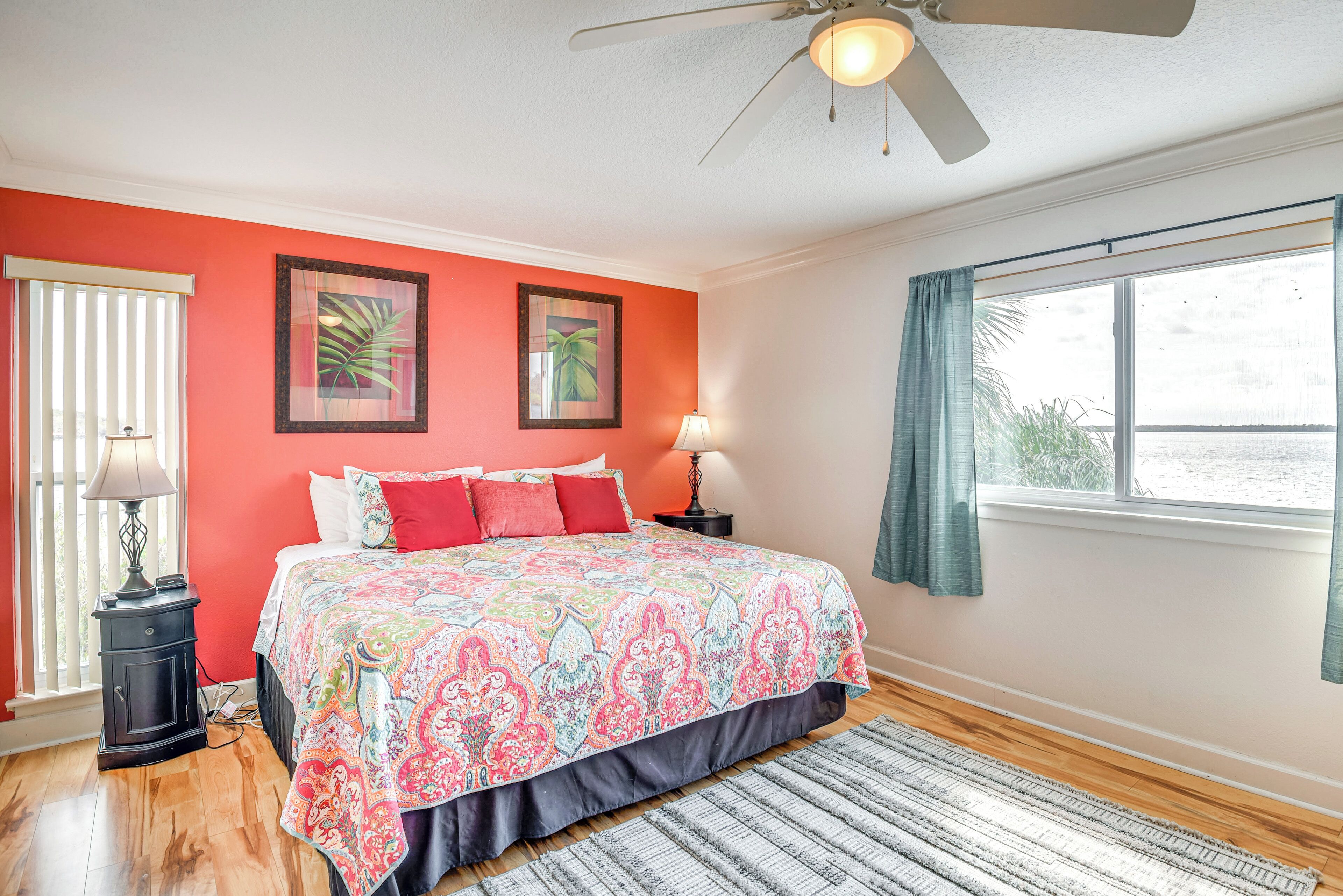 A bedroom with a large bed, a ceiling fan, a window with curtains, and a view of the outdoors.