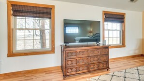 A room with two large windows with wooden frames and blinds, a flat-screen TV on a wooden dresser, and hardwood flooring.
