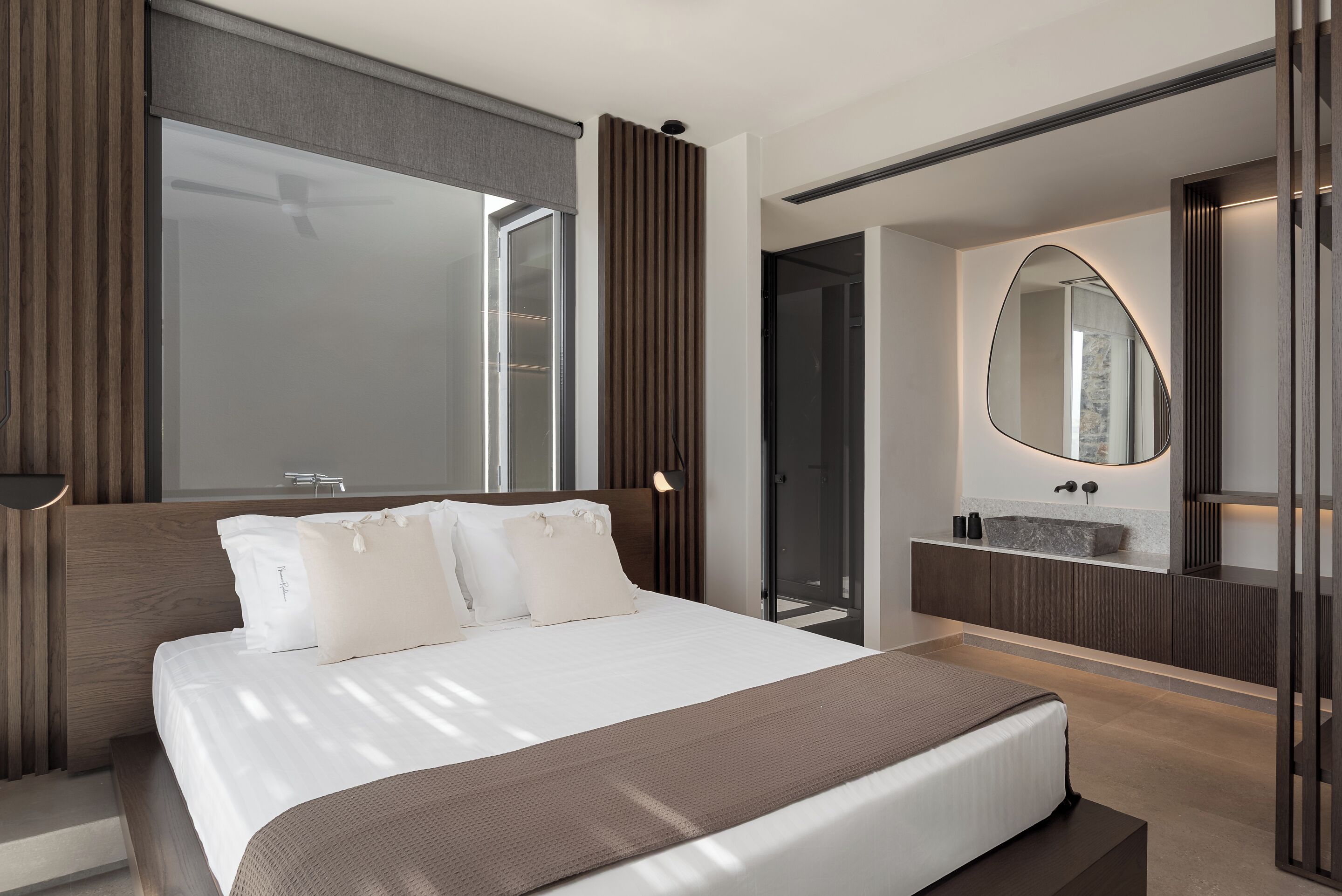 A modern hotel room with a large bed, a built-in desk, a bathroom with a vanity and mirror, and a view of the city.