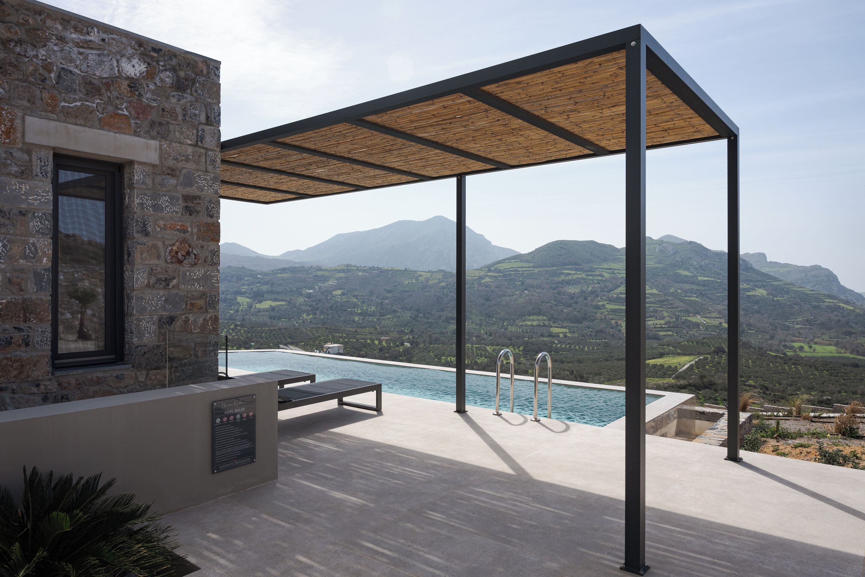 Un área de piscina moderna al aire libre con un edificio de piedra, una pérgola de madera y vistas a montañas.