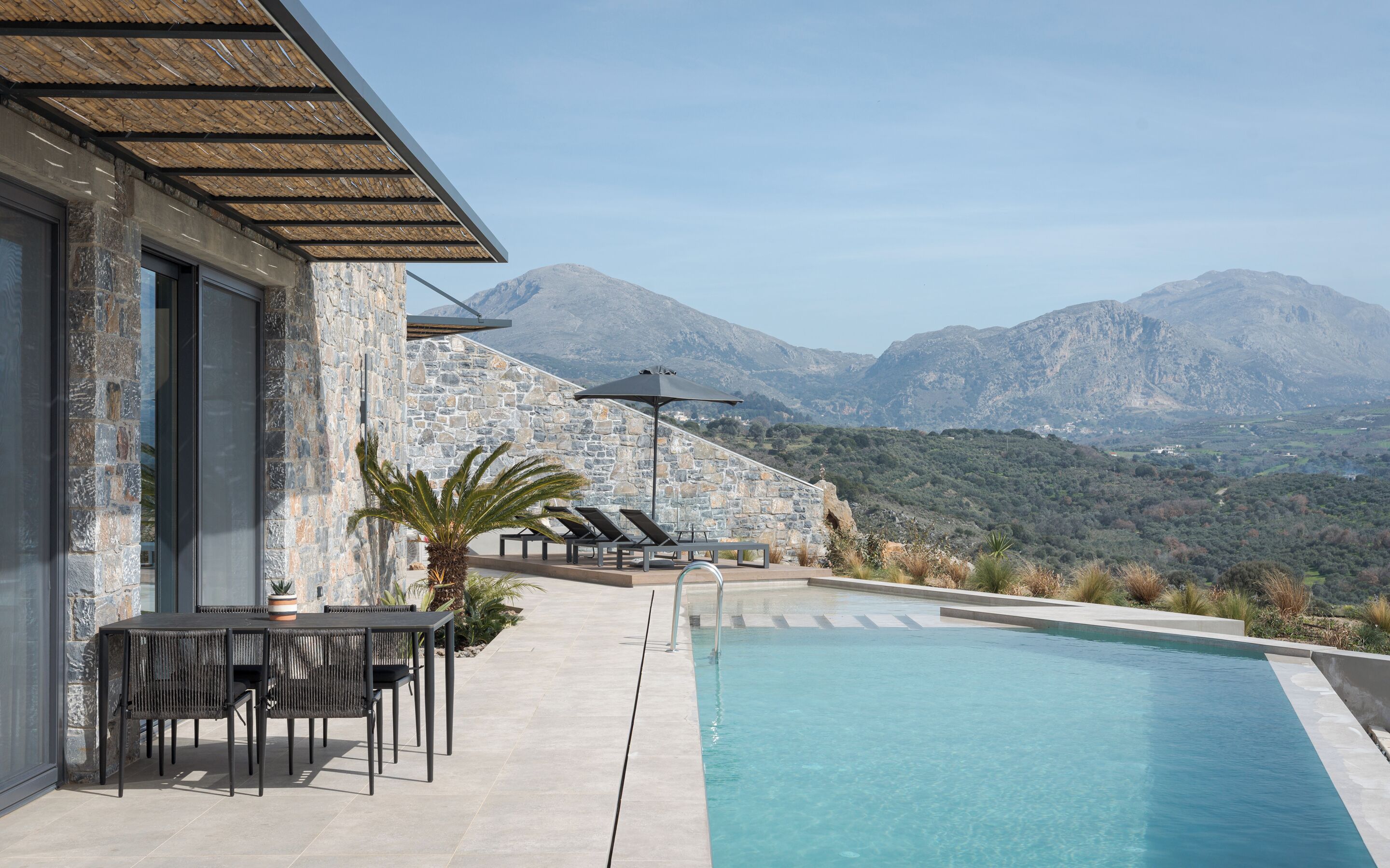 A pool area with a stone wall, lounge chairs, and a view of mountains.
