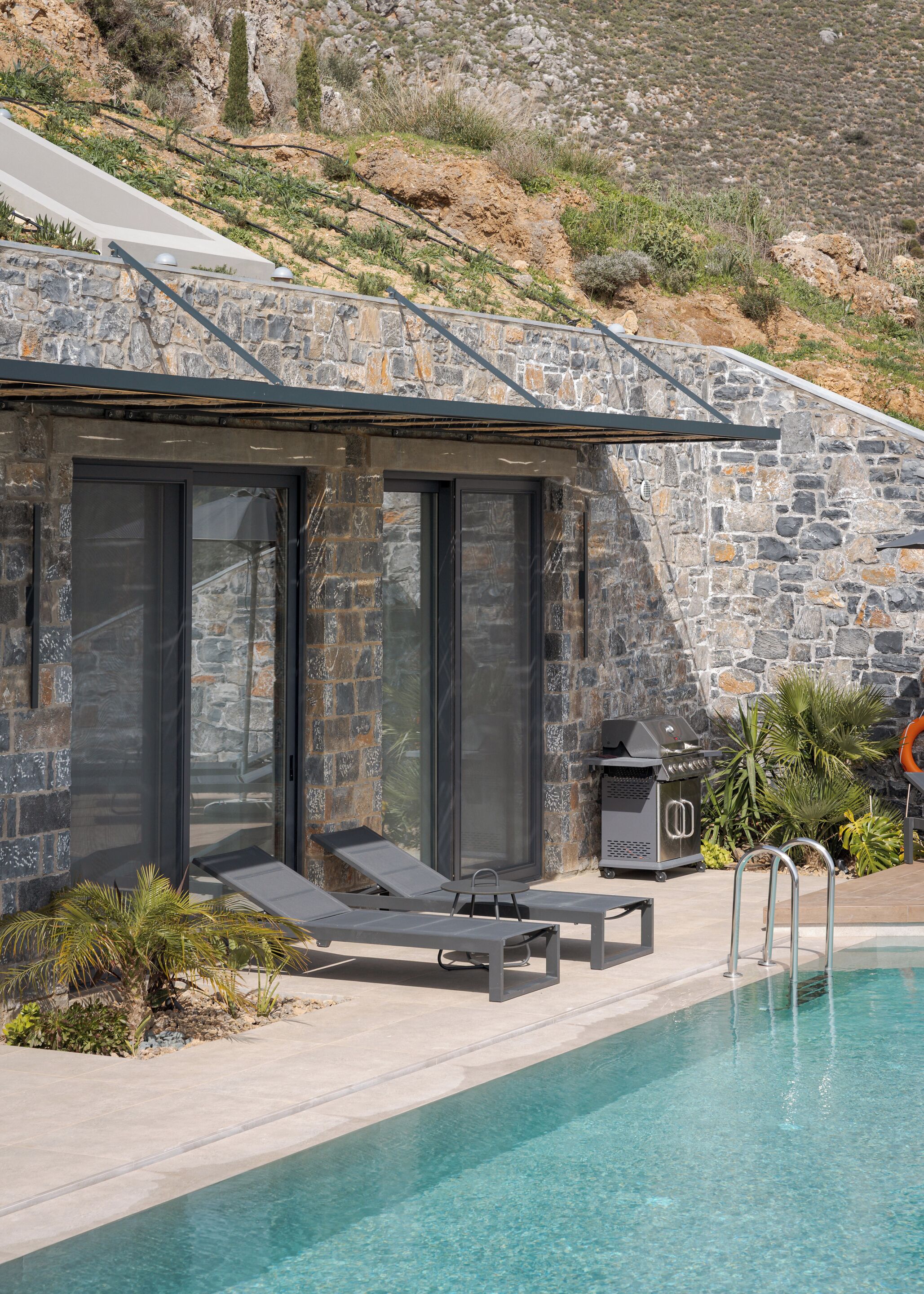 A modern outdoor pool area with stone walls, a grill, and lounge chairs.