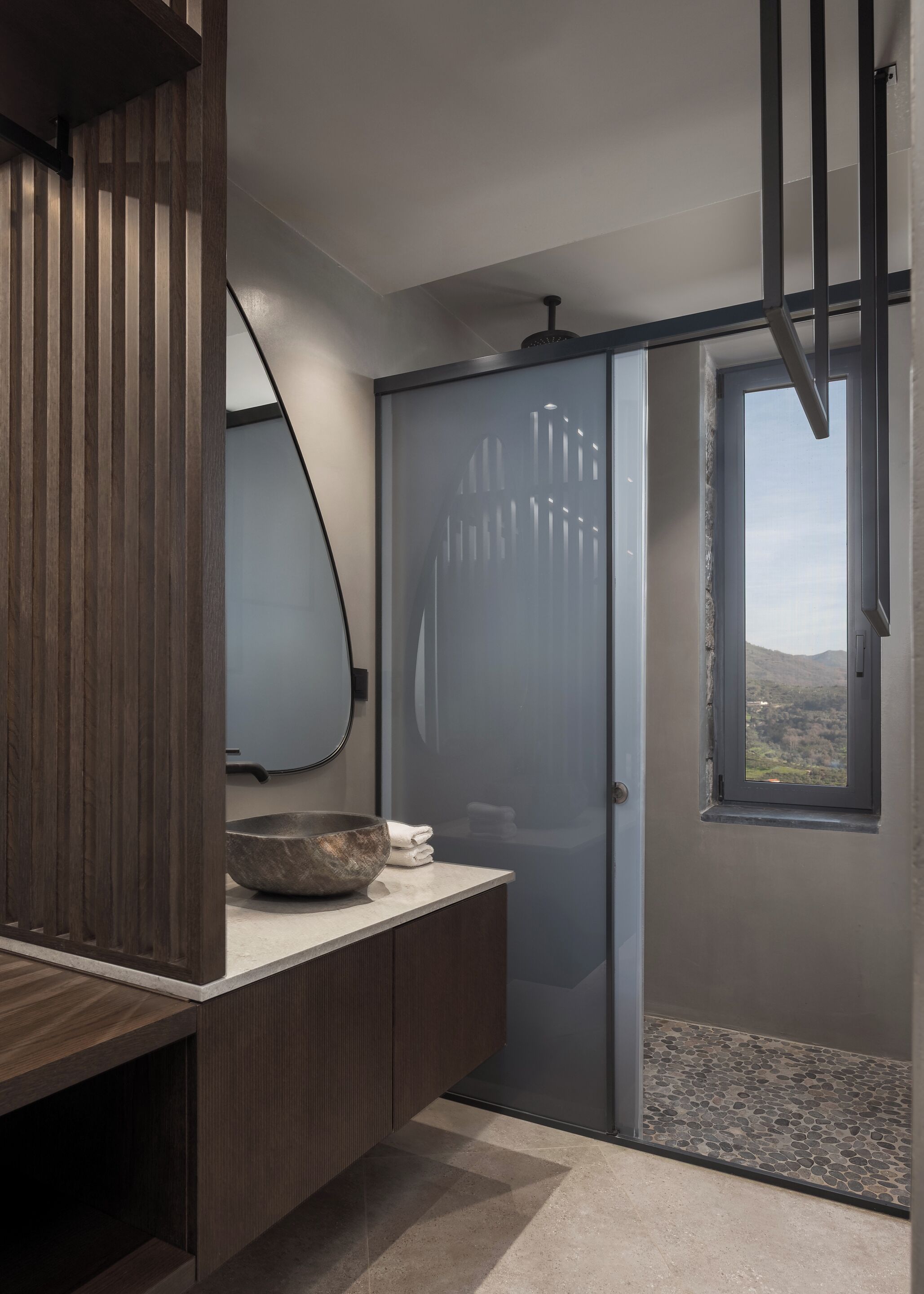 A modern bathroom with a wooden vanity, a large mirror, a glass shower enclosure, and a view of the outdoors through a window.