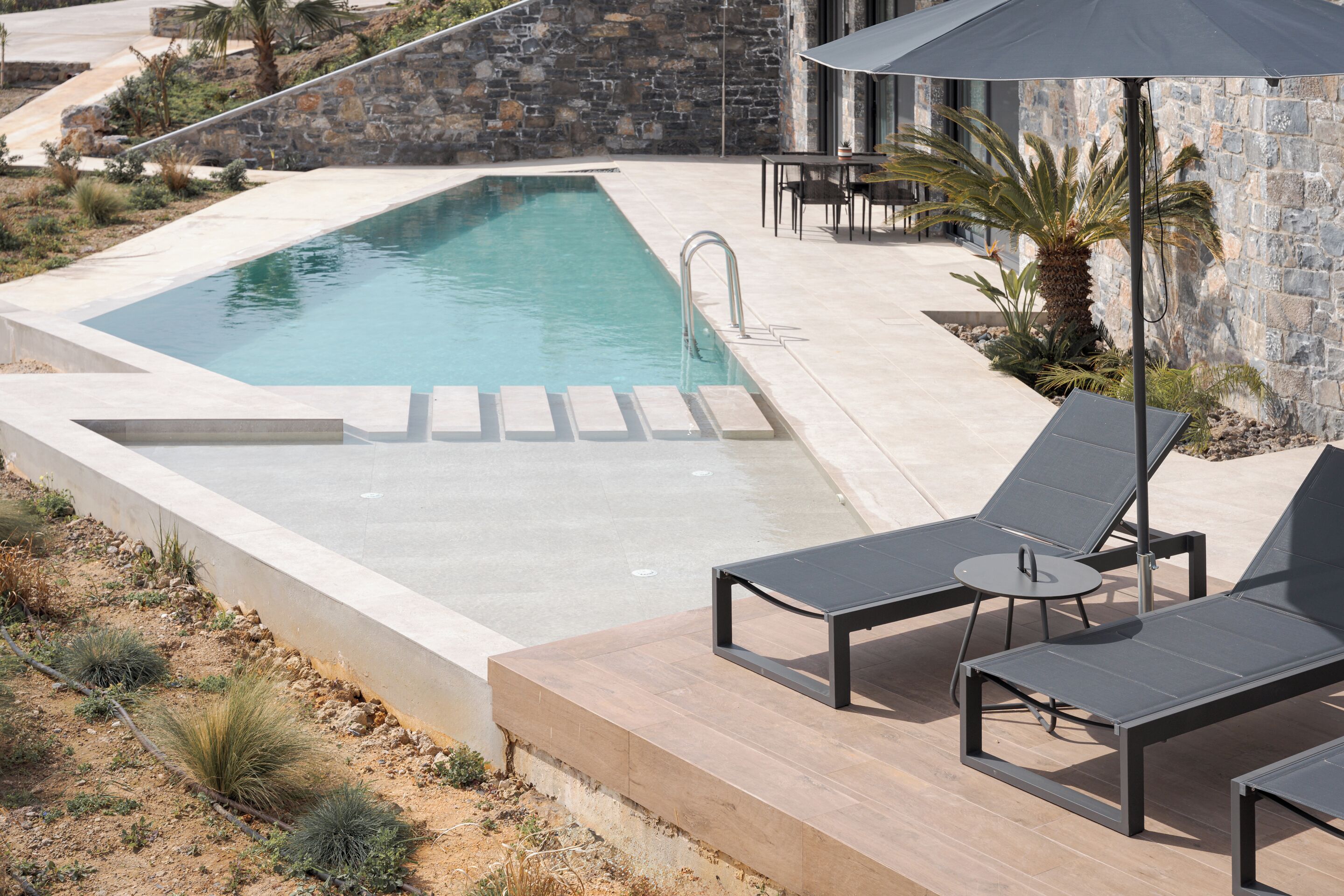 A rectangular swimming pool with a stone wall backdrop, adjacent lounge chairs, and a small table.