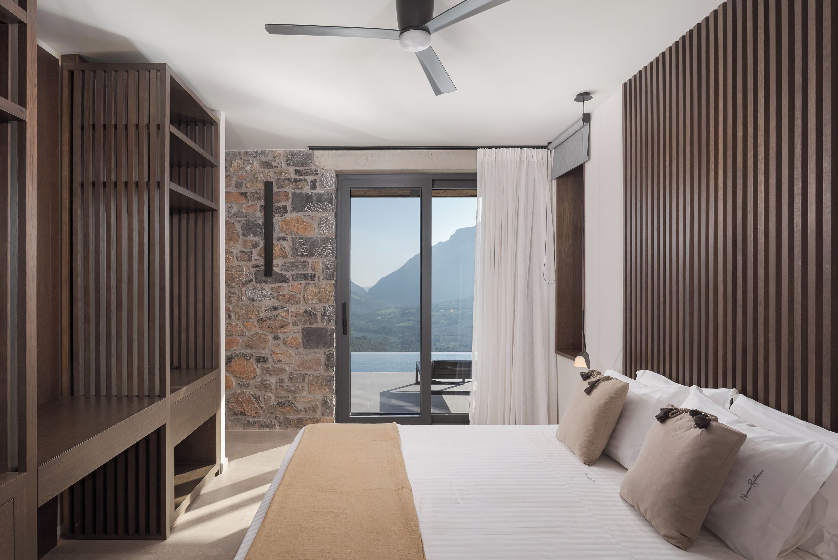 Ein Schlafzimmer mit einem Bett, Holzböden, einer Steinwand, einem Deckenventilator und Blick auf Berge.