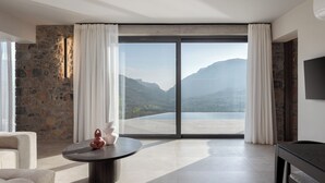 Una habitación moderna con pared de piedra, un amplio ventanal corredera que da a un balcón con vistas a la montaña, una mesa redonda con un jarrón y un televisor de pantalla plana montado en la pared.