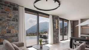 Una sala de estar moderna con pared de piedra, ventanas amplias, un sofá, una mesa de centro y un comedor con vistas a montañas.