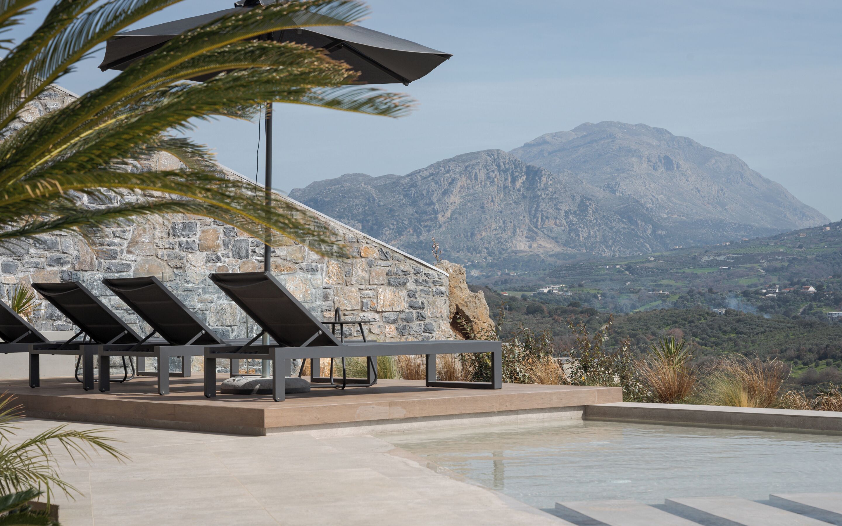 A pool area with lounge chairs and umbrellas, a stone wall, and a mountain view.