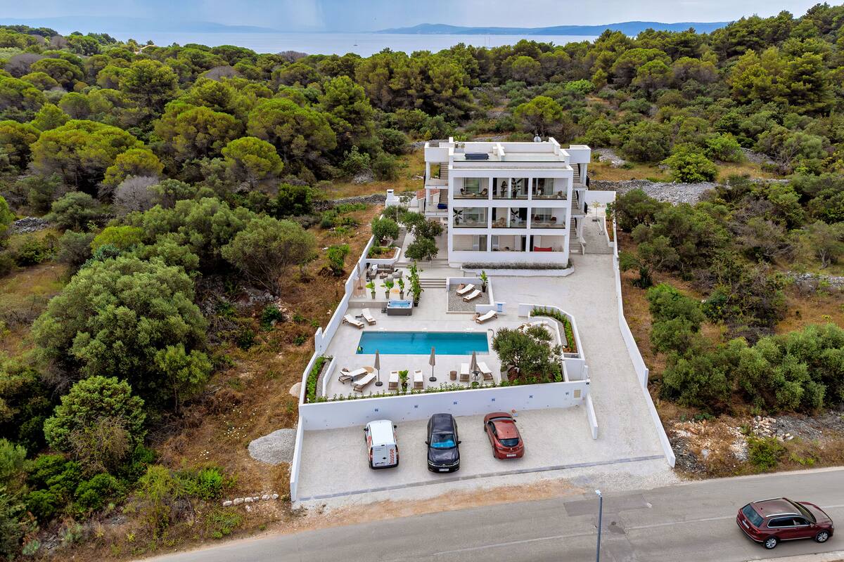 A modern two-story building with a swimming pool, surrounded by lush greenery and a road in the foreground.