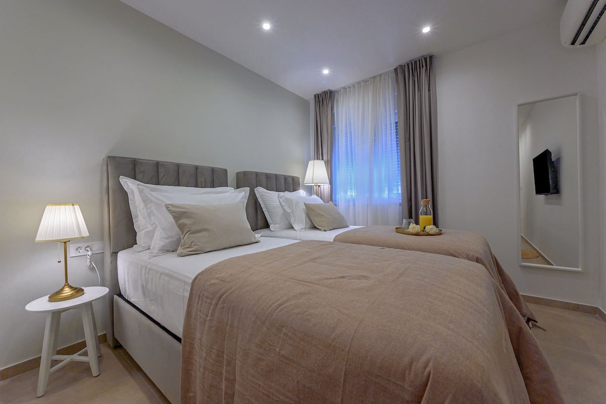 A hotel room with two beds, a bedside table with a lamp, a flat-screen TV mounted on the wall, and a window with curtains.