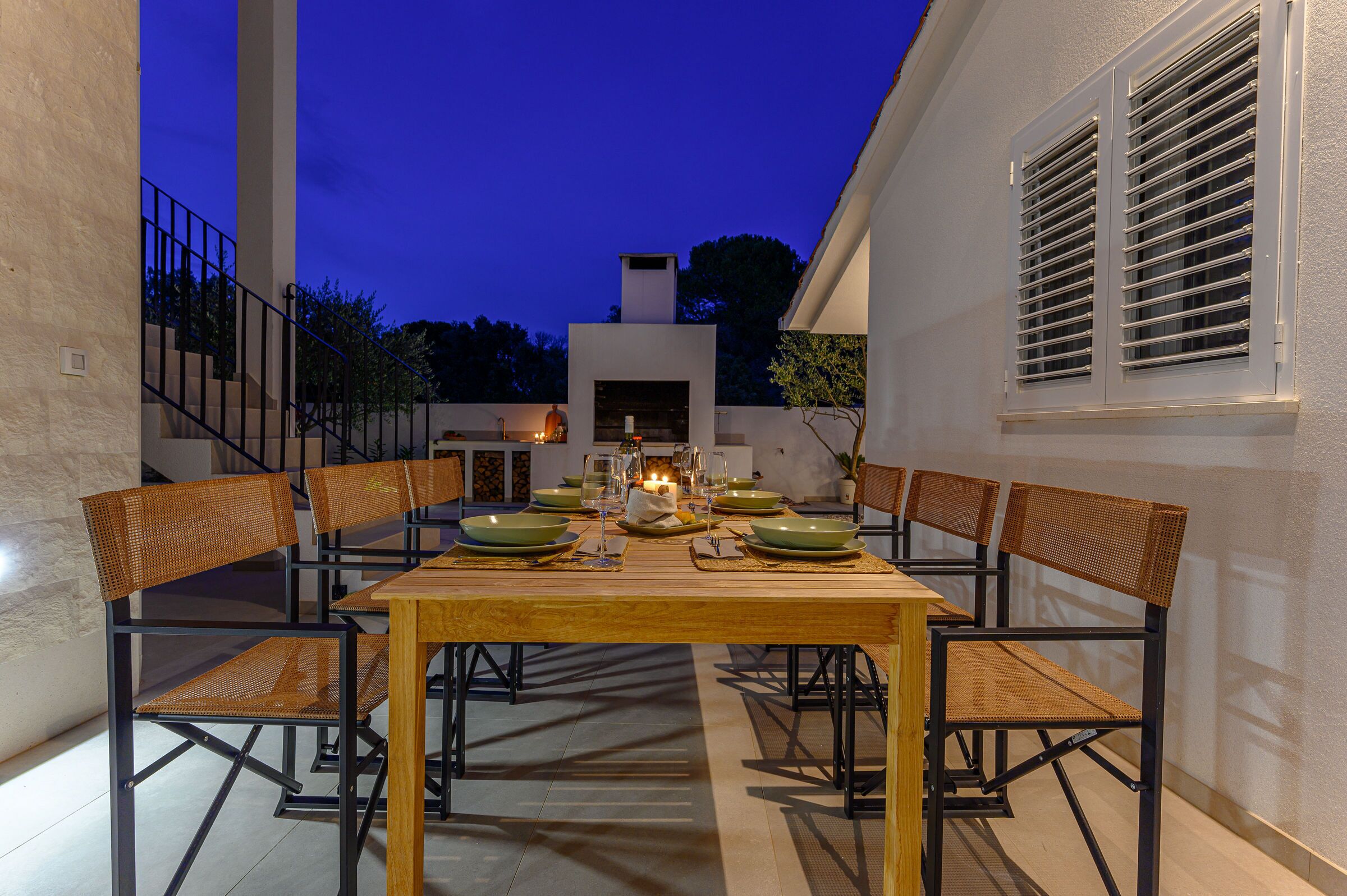 An outdoor dining area with a wooden table and chairs, surrounded by white walls and a staircase.