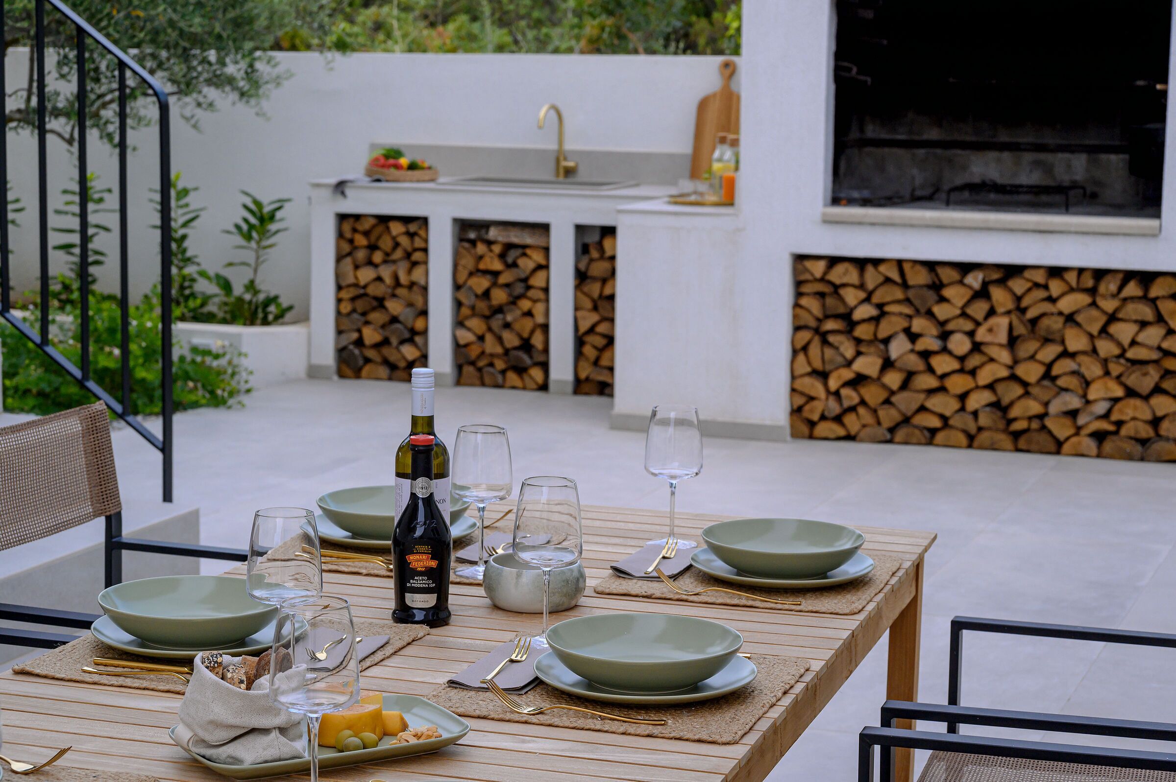 Un espace de restauration en plein air avec une table dressée pour deux personnes, une bouteille de vin et une vue sur une cuisine moderne équipée d’un évier et d’une pile de bois de chauffage.