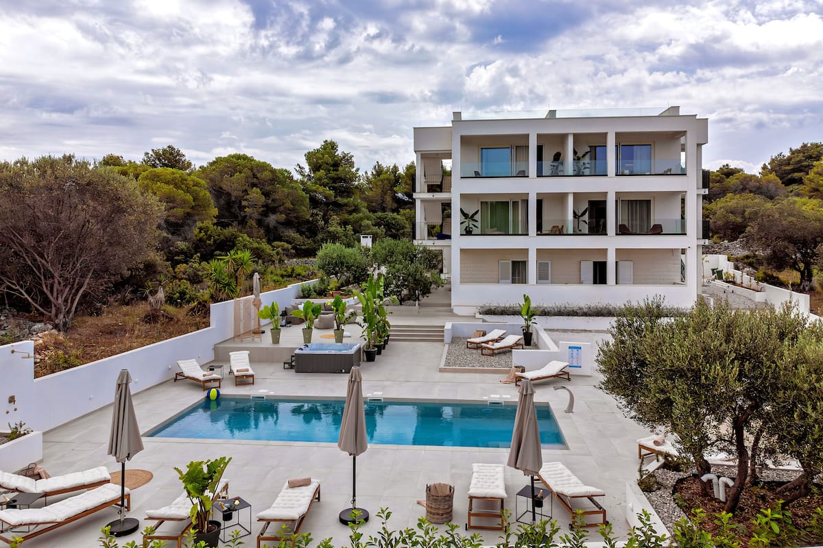 A modern building with a pool, lounge chairs, and umbrellas.