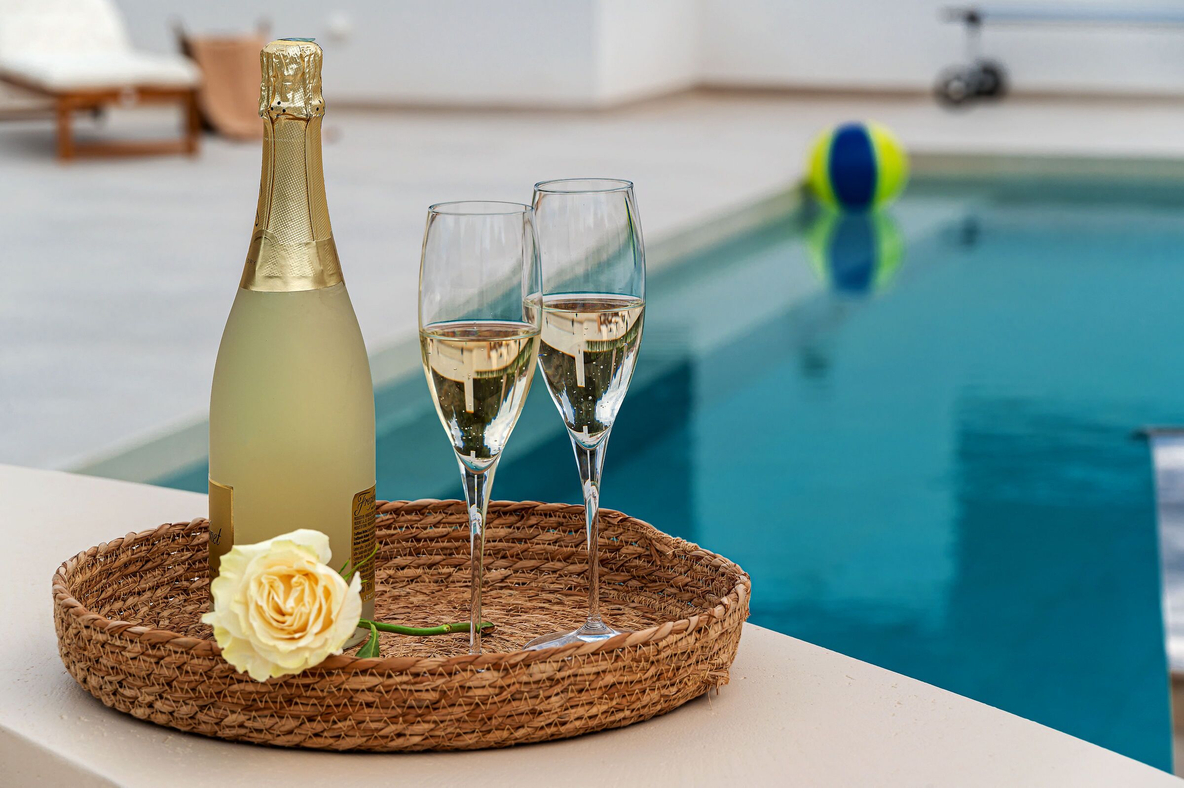 A bottle of champagne and two glasses on a woven tray with a white rose beside them, placed by a poolside.