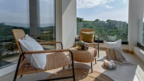Een balkon met rieten stoelen, een kleine tafel met fruit en kaarsen, en uitzicht op een bebost landschap.